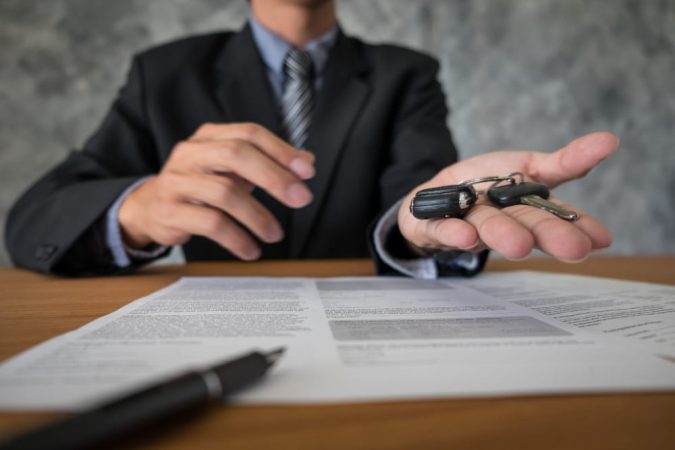 Un professionnel en costume assis à un bureau tend des clés de voiture au-dessus d'un contrat assurance automobile.
