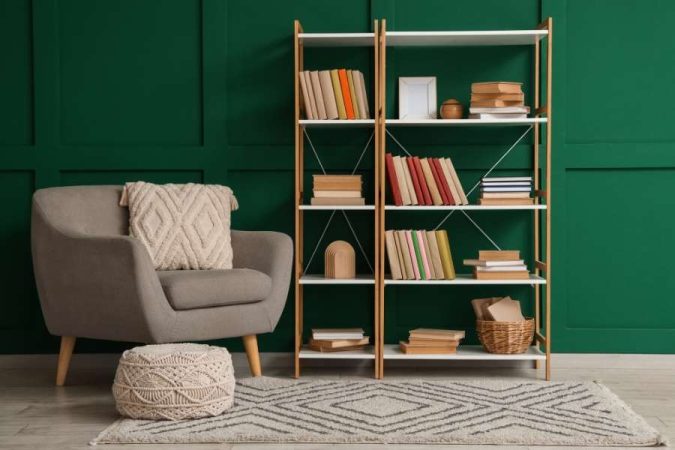 Une bibliothèque murale en bois remplie de livres, installée contre un mur vert profond près d'un fauteuil confortable.