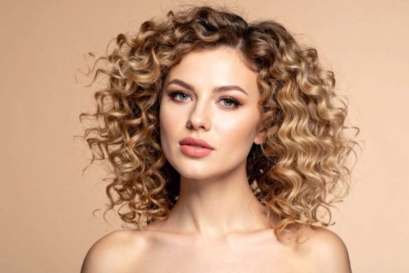 Une femme blonde aux magnifiques cheveux bouclés pose de face avec un maquillage soigné.