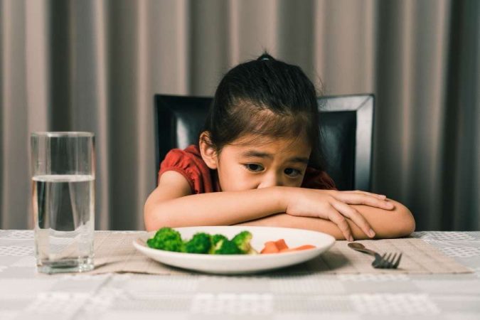 Une jeune fille boude devant son assiette de brocolis et de carottes, illustrant parfaitement le moment où un enfant refuse de manger.